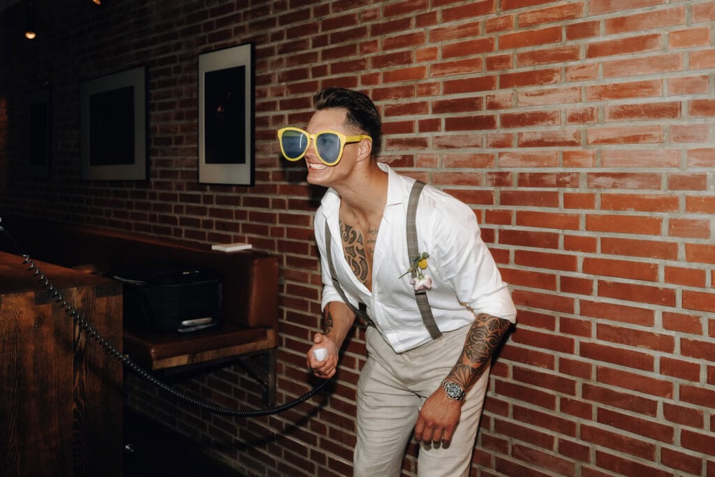 Hochzeitsgast lacht ausgelassen mit großer gelber Party-Sonnenbrille vor einer Fotobox. Hochzeit Erding Party.