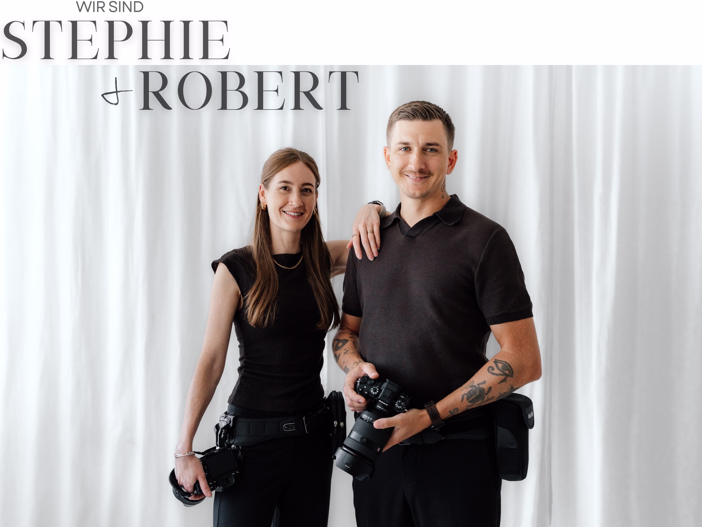 Ein Porträtfoto des Fotografen-Duos Stephie & Robert vor einem weißen Vorhang-Hintergrund, beide tragen schwarze Kleidung und halten professionelle Kameras.